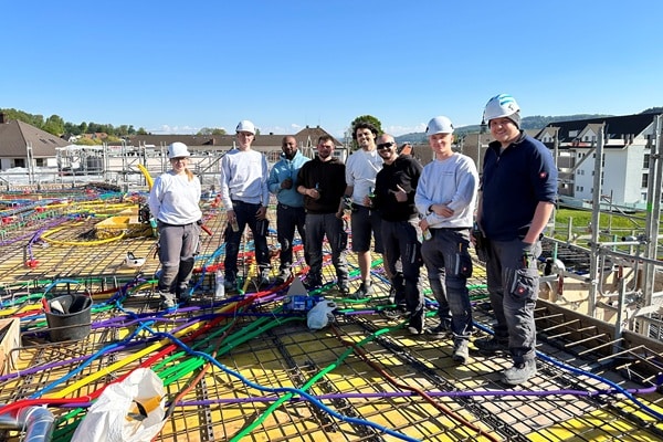 Ein Team von Elektroinstallateuren steht auf einer Rohbaustelle vor einer Bodenplatte mit verlegten farbigen Elektro-Leerrohren. Die Aufnahme zeigt die fachgerechte Elektro-Rohinstallation vor dem Betonieren und unterstreicht Teamarbeit sowie präzise Ausführung im modernen Wohnungsbau